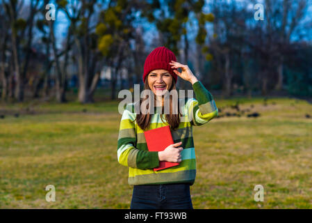 Teenager-Mädchen im roten Hut und grün gestreifte Pullover lacht Andholding Buch mit rotem Einband stehen auf einer Grünfläche im Park. Stockfoto