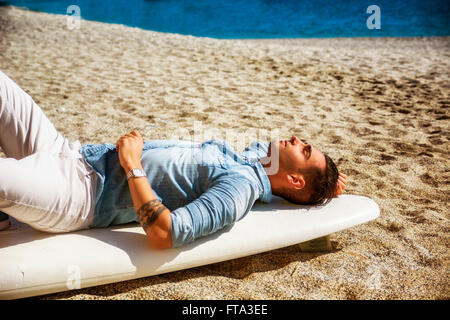 Stilvolle hübscher junger Mann Handauflegen Surfbrett am Strand in der Sonne, mit geschlossenen Augen, die Wellen warten ruhen Stockfoto