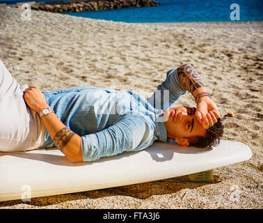 Stilvolle hübscher junger Mann Handauflegen Surfbrett am Strand in der Sonne Stockfoto