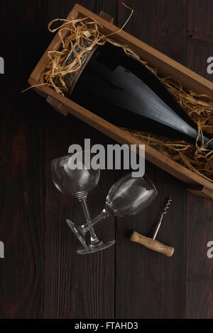 Flasche Rotwein in Holzkiste gefüllt mit Stroh neben, leere Gläser mit Korkenzieher auf Holztisch. Flache Lay, oben vi Stockfoto