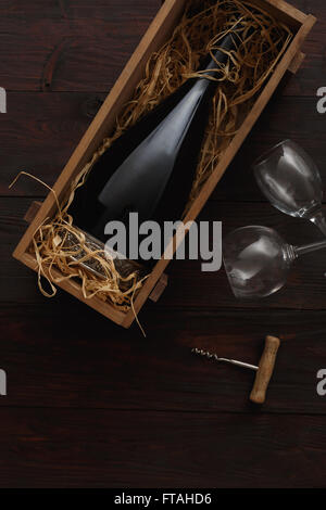 Flasche Rotwein in Holzkiste gefüllt mit Stroh neben, leere Gläser mit Korkenzieher auf Holztisch. Flache Lay, oben vi Stockfoto