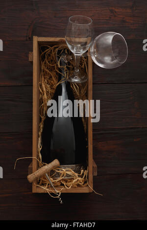 Flasche Rotwein in Holzkiste gefüllt mit Stroh neben, leere Gläser mit Korkenzieher auf Holztisch. Flache Lay, oben vi Stockfoto