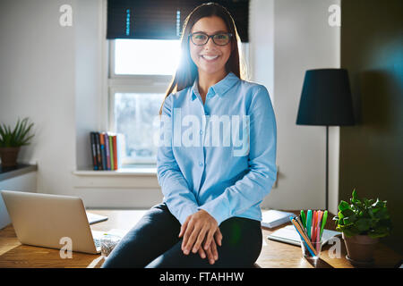 Entspannt, zuversichtlich junge Unternehmerin oder Unternehmer mit Brille sitzt auf der Kante von ihrem Schreibtisch in eine home-Office lächelnd ein Stockfoto