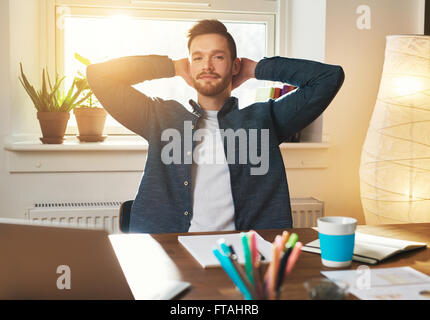 Entspannte Unternehmer sitzen mit seinem Handzeichen hinter seinen Hals mit einem schweren spekulativen Ausdruck in die Kamera schaut, Stockfoto