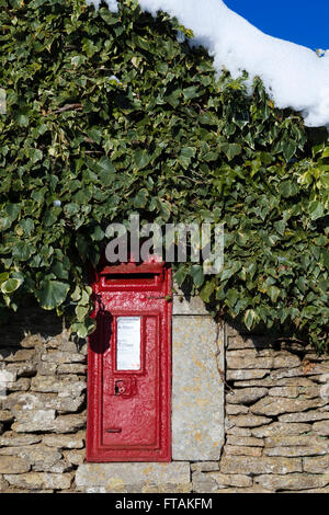 Ein tiefes roter traditionellen Briefkasten inmitten einer Trockensteinmauer in den Cotswolds, Gloucestershire Uk, Efeu und mit einer Schicht aus Schnee an der Spitze in diesem Winter Landschaft Szene abgedeckt. Stockfoto