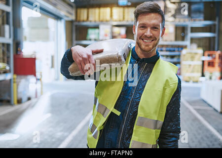 Schließen Sie jungen Mann arbeitet in einem Lager steht mit einem Beutel des Produkts über die Schulter grinsend fröhlich in die Kamera Lächeln, Stockfoto