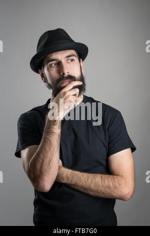 Denken Hipster trägt schwarze T-shirt und Hut streicheln Bart wegsehen. Kopfschuss-Porträt über graue Studio-Hintergrund Stockfoto