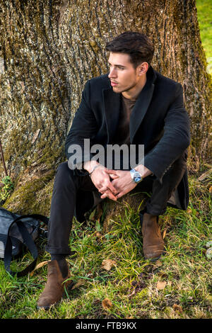 Hübscher junger Mann lehnte sich gegen Baum, Blick in die Kamera, an einem sonnigen Tag trägt einen schwarzen Mantel Stockfoto