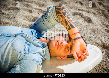 Stilvolle hübscher junger Mann Handauflegen Surfbrett am Strand in der Sonne Stockfoto