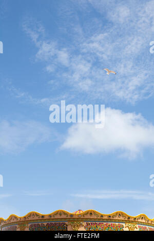 Ein Segull fliegt über einen Kreisverkehr, bunt bemalt, an einem Strand. Mit blauem Himmel und Sonnenschein ist die Szene eines heißes Wetter und Sommerferien. Stockfoto