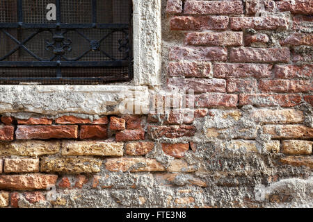 Bunt bröckelnde, freiliegende und verfallende Ziegelarbeiten um ein mit Steinen umrahmtes, kunstvoll versperrtes Fenster in einem Gebäude in Venedig Stockfoto