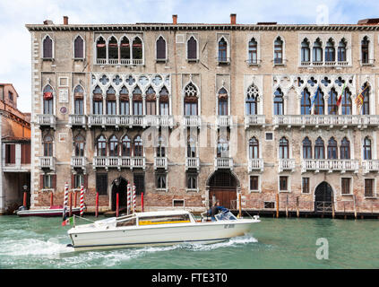 Klassisches Hafengebäude mit verzierten Fenstern und Balkonen mit roten Anlegestellen und einer vorbeifahrenden Bootsanlegestelle auf dem Canal Grande, Venedig, Italien Stockfoto