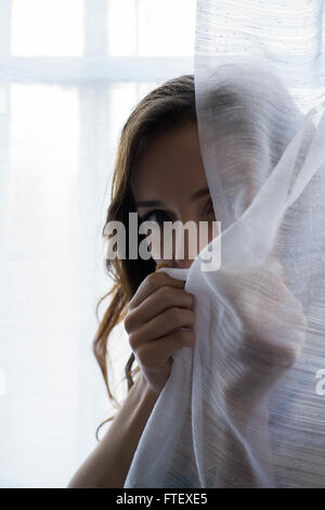 Ernste junge Frau versteckt sich hinter die Vorhänge am Fenster Stockfoto