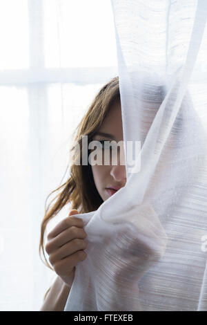 Ernste junge Frau versteckt sich hinter die Vorhänge am Fenster Stockfoto