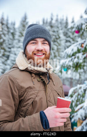 Glückliche junge bärtigen Mann trinken heißen Kaffee in der Nähe von geschmückter Weihnachtsbaum im Winterwald Stockfoto