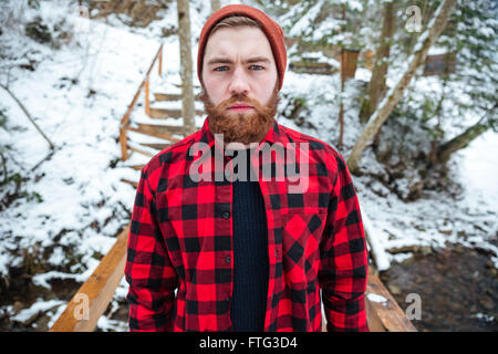Porträt von hübscher ernst junger Mann mit Bart in Hut und Plaid Shirt im Winterwald Stockfoto