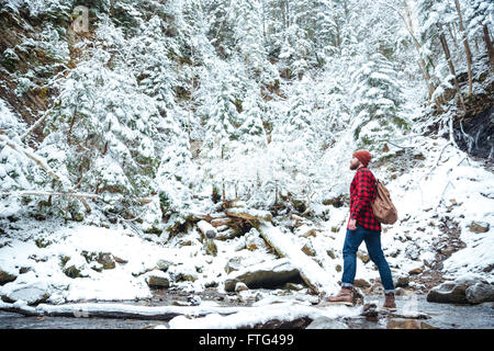 Hübscher bärtiger junger Mann im karierten Hemd mit Rucksack zu Fuß in der Nähe des Flusses im Winterwald Stockfoto