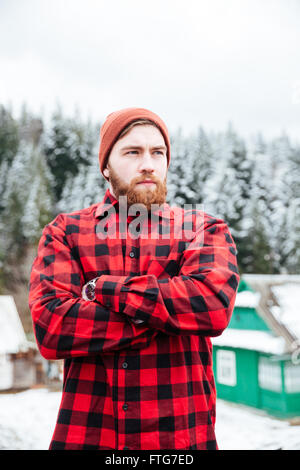 Porträt der bärtige junge Mann roten Hut und kariertes Hemd mit Armen stehend überquerte im Winterdorf in der Nähe von Wald Stockfoto