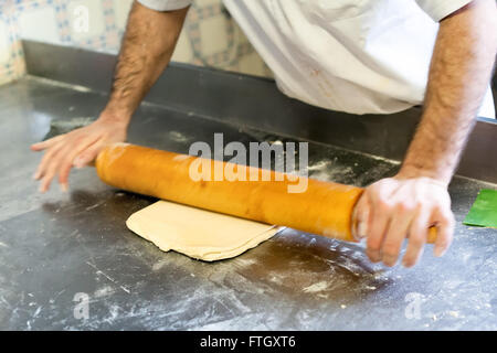 Close Up Aktion erschossen von männlichen Baker ausrollen Teig mit hölzernen Nudelholz auf Metalloberfläche inmitten der professionellen Bäckerei Stockfoto