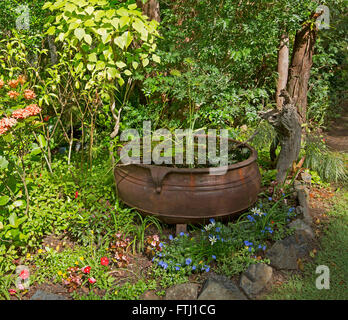 Spektakulärer Garten mit großen rostigen Kessel Wasserspiel mit rot, blau, weiße Blumen & Dichte Smaragd grüne Sträucher hinter Stockfoto