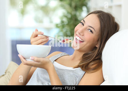 Glückliches Mädchen essen Müsli mit Obst aus einer Schüssel sitzt auf einer Couch zu Hause Stockfoto