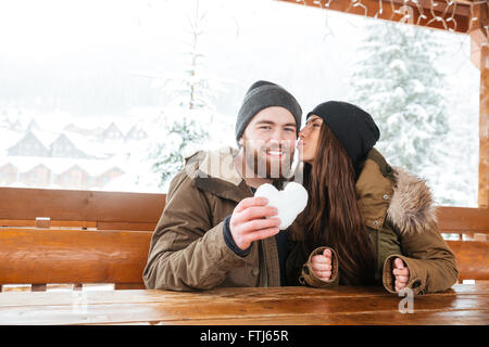 Glückliches junges Paar küssen im Chalet Terrasse und hält Herz aus Schnee im Winter gemacht Stockfoto
