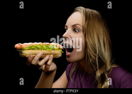 Junge hübsche Mädchen essen einen chaotischen Hotdog auf schwarzem Hintergrund isoliert Stockfoto