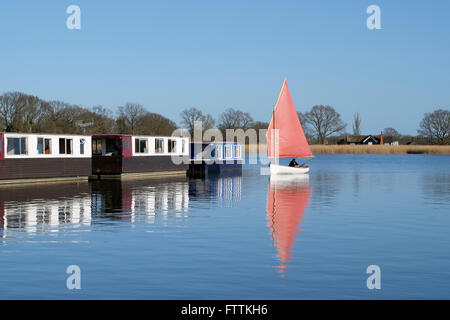 Eine traditionelle Klinker geschält Segeln Jolle Segeln vorbei an einer Reihe von Hausbooten auf Hickling Broad, Norfolk, England, UK Stockfoto