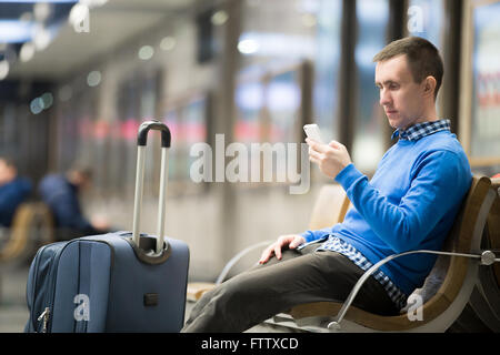 Porträt von schönen jungen Mann casual-Style Kleidung sitzen auf dem Sitz in modernen Flughafen mit Smartphone. PKW Stockfoto