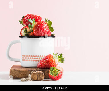 Frische reife Erdbeeren im Land Stil Emaille Becher auf rustikalem Holzbrett über rosa Pastell Hintergrund Stockfoto