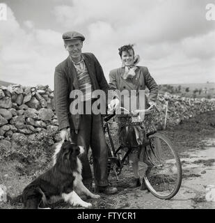 Die 1950er Jahre, historisches Bild von J Allan Cash aus dem Westen Irlands, lächeln alle als irischer Bauer mit Schafhund und seine Frau mit ihrem Radstopp auf einer Landstraße für ihr Foto. Stockfoto