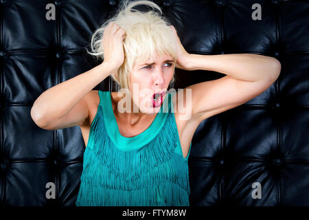 blonde Frau im grünen Kleid schreien vor einer Wand aus schwarzem Leder Stockfoto