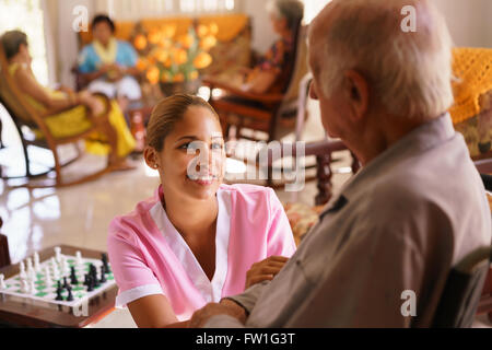 Alte Menschen in geriatrischen Hospiz: junge attraktive hispanic Frau arbeitet als Krankenschwester kümmert sich ein senior Mann im Rollstuhl. Stockfoto