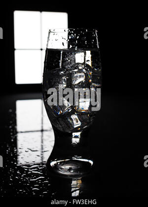 Ein Glas mit Eiswürfeln gegen das Fenster in der Dunkelkammer-Stillleben Stockfoto