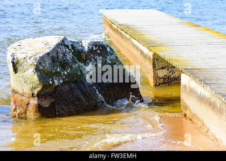 Einen großen Granitfelsen neben einem hölzernen und konkreten Pier. Die Pier hat einen Kanal Wasser neben dem Stein. Stockfoto