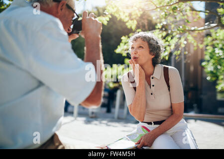Schöne ältere Frau bläst einen Kuss und Kamera im freien sitzend auf einer Bank in der Stadt. Frau von seinem h fotografiert Stockfoto