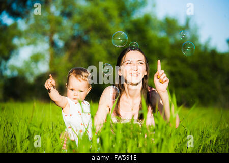 Mama und Baby Mädchen Verlegung auf Rasen und fröhlich fangen Seifenblasen mit Fingern. Glückliche Familien-Konzept. Stockfoto