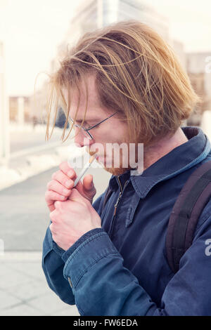 junger Mann mit roten langen Haaren und Brille, eine Zigarette rauchend Stockfoto