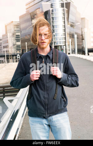 junger Mann mit langen roten Haaren, Bart und Brille, die auf der Straße mit einem Rucksack Stockfoto