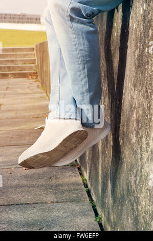 Nahaufnahme von Mann in Jeans und Turnschuhen auf einer Mauer sitzend Stockfoto