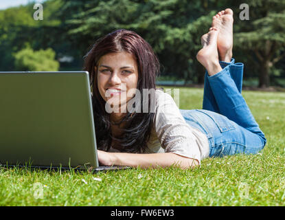 Junge Brünette Frau mit Laptop-Computer, auf dem Rasen in einem Park liegen Stockfoto