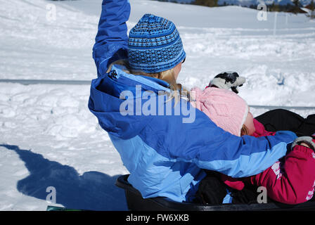 Blonde behaarte Mutter in blaue Skijacke mit Tochter von hinten nach unten im Schnee Rodeln. Stockfoto