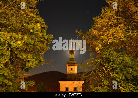 Kirche und Laub von Straßenlaternen mit dem Mond hinter dem Kirchturm beleuchtet. Loket, Bezirk Sokolov, Böhmen, Tschechische Republik. Stockfoto