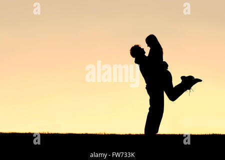 Eine Silhouette ein glückliches junges Paar umarmt, mit der Mann die Frau Weg von ihren Füßen, heben, bei Sonnenuntergang.  Himmel lässt Raum für co Stockfoto