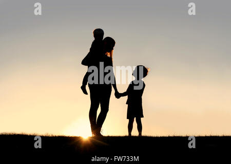 Silhouette einer jungen Mutter liebevoll Hand in Hand mit ihr glückliches Kind mit gedrückter Bruder Baby außerhalb bei Sonnenuntergang Stockfoto