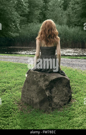 Rückansicht einer jungen Frau im gepunkteten Kleid sitzt auf einem Felsen Stockfoto