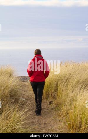 Einsame Frau zu Fuß zum Strand durch Dünengebieten Grass. Allonby, Cumbria, England, Vereinigtes Königreich, Europa. Stockfoto