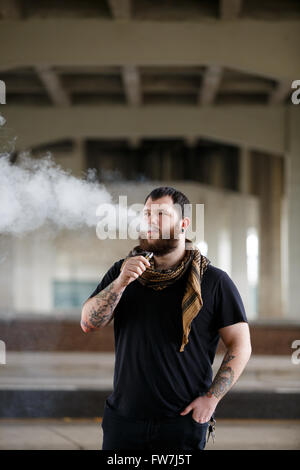 EUGENE, OR - 20. März 2016: Urban Porträt eines bärtigen Hipster mit Tätowierungen Dampfen mit einem benutzerdefinierten mod Vape-Gerät. Stockfoto