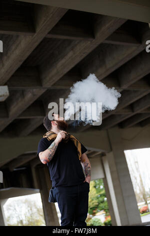 EUGENE, OR - 20. März 2016: Urban Porträt eines bärtigen Hipster mit Tätowierungen Dampfen mit einem benutzerdefinierten mod Vape-Gerät. Stockfoto