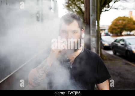 EUGENE, OR - 20. März 2016: Urban Porträt eines bärtigen Hipster mit Tätowierungen Dampfen mit einem benutzerdefinierten mod Vape-Gerät. Stockfoto